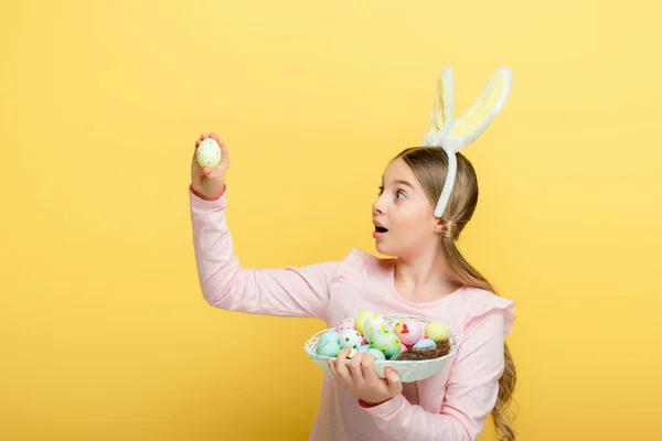 Enfant surpris avec des oreilles de lapin regardant l'oeuf de Pâques isolé sur jaune — Photo de stock