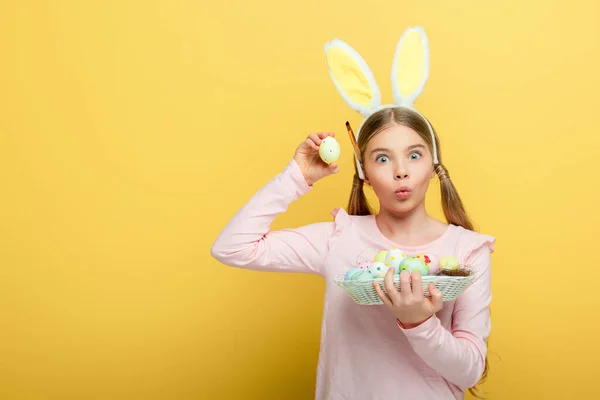 Schockiertes Kind mit Hasenohren blickt in die Kamera und hält Ostereier isoliert auf gelb — Stockfoto