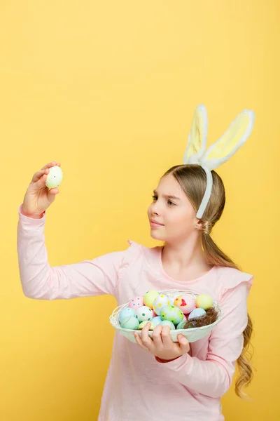 Милый ребенок с кроличьими ушами глядя на пасхальное яйцо изолированы на желтый — стоковое фото