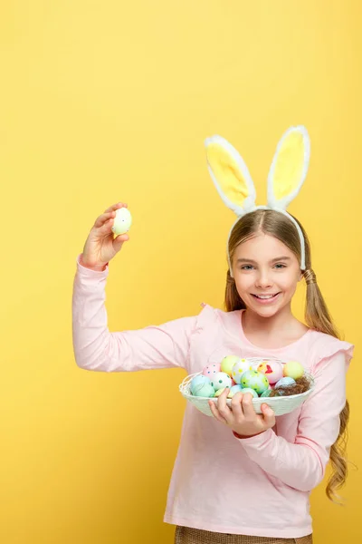 Enfant heureux avec des oreilles de lapin tenant des œufs de Pâques dans le panier isolé sur jaune — Photo de stock