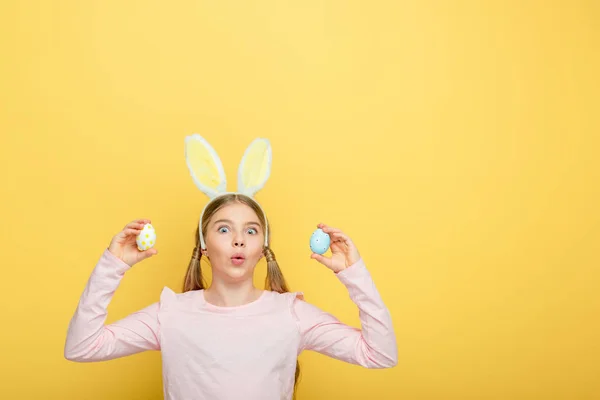 Shocked child with bunny ears holding painted easter eggs isolated on yellow — Stock Photo