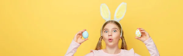 Plan panoramique de l'enfant choqué avec des oreilles de lapin tenant des œufs de Pâques peints isolés sur jaune — Photo de stock