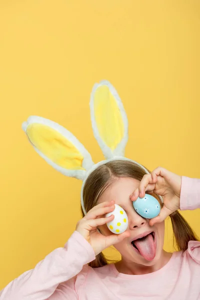 Enfant avec des oreilles de lapin collant la langue et couvrant les yeux avec des œufs de Pâques isolés sur jaune — Photo de stock