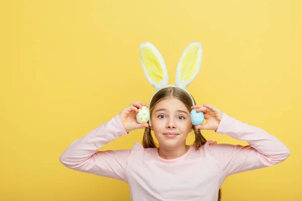 Mignon enfant avec des oreilles de lapin tenant des œufs de Pâques peints isolés sur jaune — Photo de stock
