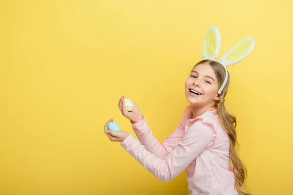 Positive child with bunny ears holding painted easter eggs isolated on yellow — Stock Photo