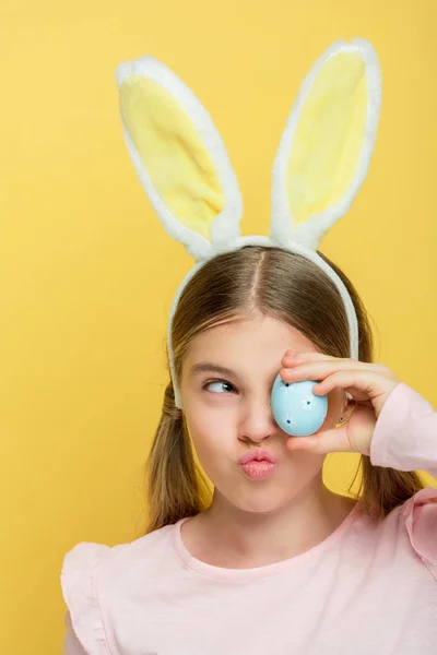 Divertido niño con orejas de conejo que cubre el ojo con huevos de Pascua punteados aislados en amarillo - foto de stock