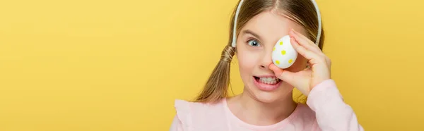 Plano panorámico de niño emocional con orejas de conejo que cubre el ojo con huevos de Pascua punteados aislados en amarillo - foto de stock