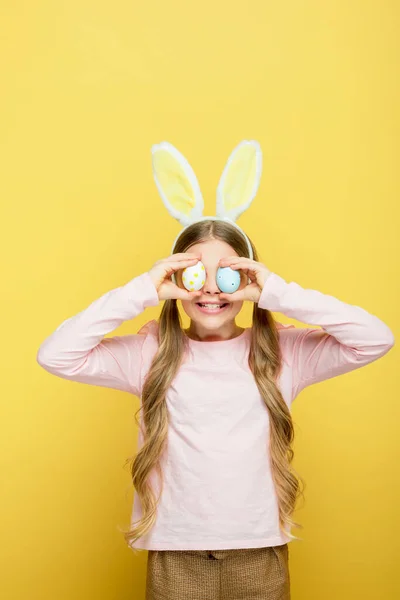 Весёлый ребёнок с кроличьими ушами, закрывающими глаза пасхальными яйцами, изолированными на жёлтом — стоковое фото