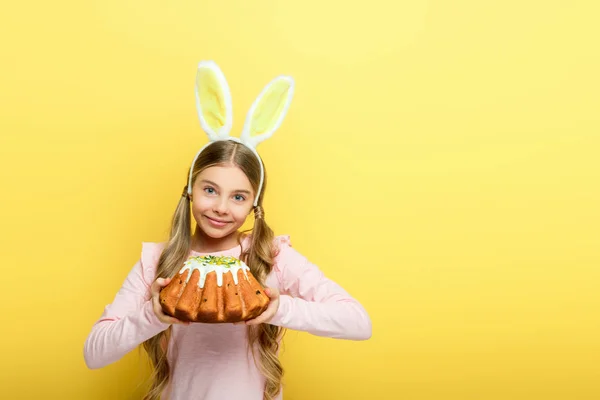 Criança feliz com orelhas de coelho segurando bolo de Páscoa isolado no amarelo — Fotografia de Stock
