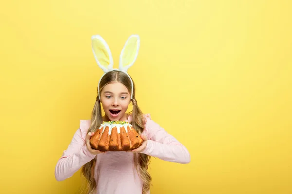 Шокированный ребенок с кроличьими ушами, держащий пасхальный торт изолирован на желтый — стоковое фото