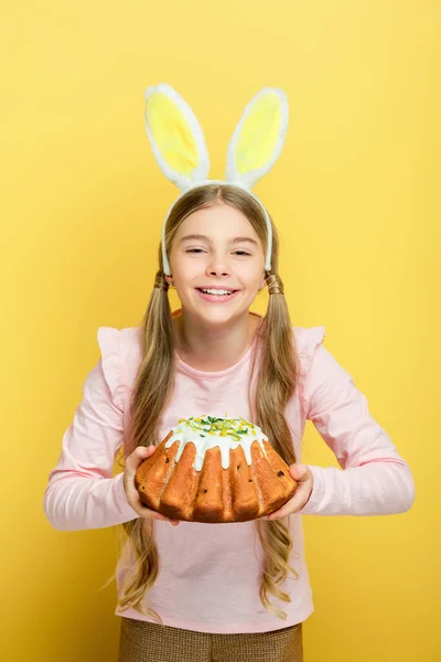 Enfant souriant avec des oreilles de lapin tenant gâteau de Pâques isolé sur jaune — Photo de stock