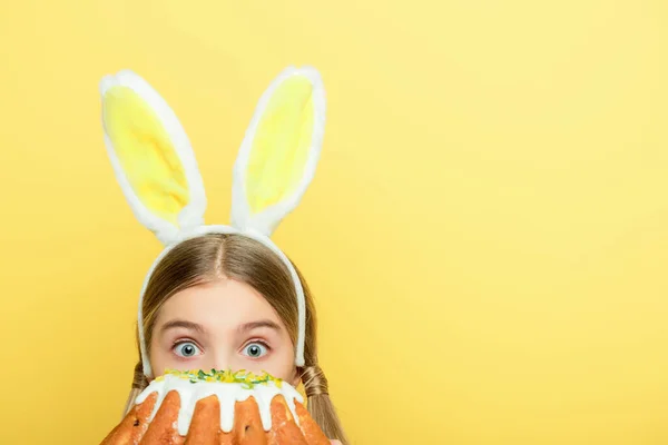 Chaval con orejas de conejo cubriendo la cara con pastel de Pascua aislado en amarillo - foto de stock