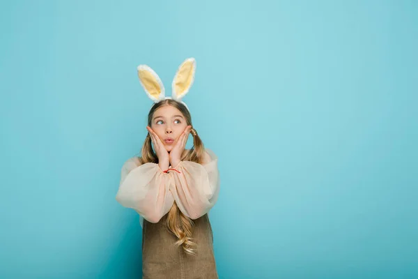 Enfant surpris avec des oreilles de lapin touchant le visage et regardant loin isolé sur bleu — Photo de stock