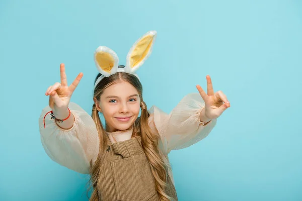 Fröhliches Kind mit Hasenohren zeigt Friedenszeichen isoliert auf blauem Grund — Stockfoto