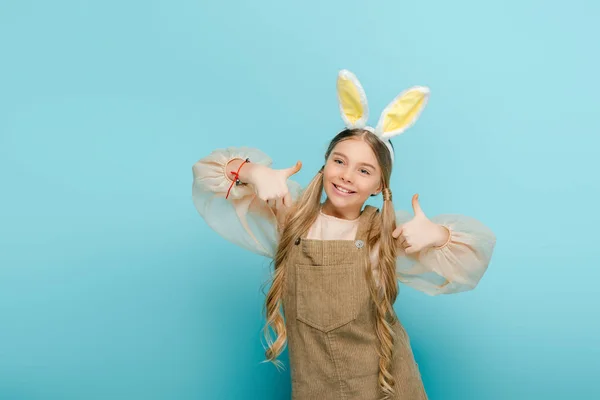 Enfant gai avec des oreilles de lapin montrant pouces isolés sur bleu — Photo de stock
