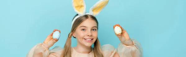 Tiro panorâmico de criança feliz com orelhas de coelho segurando ovos de páscoa pintados isolados em azul — Fotografia de Stock
