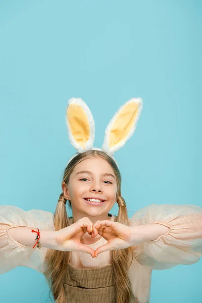 Положительный ребенок с кроличьими ушами показывая сердце с руками изолированы на синий — стоковое фото
