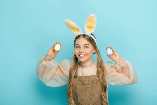 Позитивный ребенок с кроличьими ушами, держащий окрашенные пасхальные яйца на голубом — стоковое фото