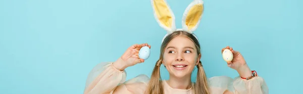 Панорамный снимок веселого ребенка с кроличьими ушами, держащего окрашенные пасхальные яйца и смотрящего в сторону изолированного на голубом — стоковое фото