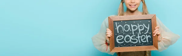 Plano panorámico de niño alegre sosteniendo pizarra con letras de Pascua feliz aislado en azul - foto de stock