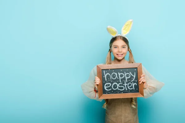 Niño alegre con orejas de conejo sosteniendo pizarra con letras de Pascua feliz en azul - foto de stock