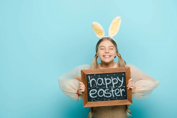Smiling kid with bunny ears holding chalkboard with happy easter lettering on blue — Stock Photo