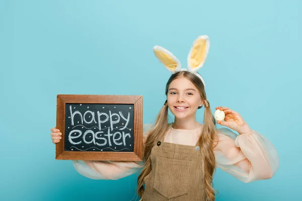 Enfant souriant avec des oreilles de lapin tenant tableau noir avec joyeuse Pâques lettrage et oeuf de poulet peint isolé sur bleu — Photo de stock