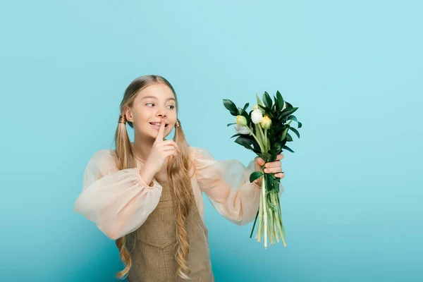 Bambino felice guardando bouquet di tulipani e mostrando il segno del silenzio isolato sul blu — Foto stock