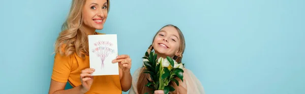 Plan panoramique de mère heureuse tenant carte de vœux avec joyeux lettrage de la fête des mères près de fille isolée sur bleu — Photo de stock