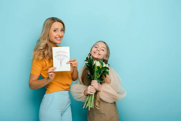 Happy mother holding greeting card with happy mothers day lettering near daughter on blue — Stock Photo