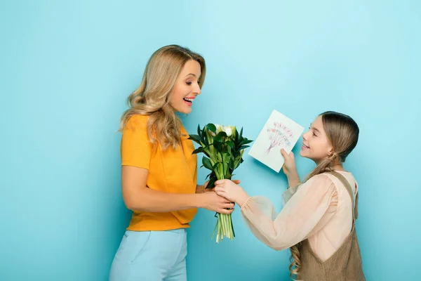 Feliz madre tomando tarjeta de felicitación con feliz día de las madres letras y flores de la hija en azul - foto de stock