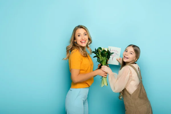 Cheerful mother taking greeting card with happy mothers day lettering and flowers from daughter on blue — Stock Photo