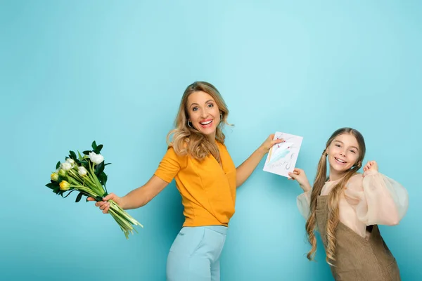 Niedliche Tochter hält Grußkarte mit 8 März Schriftzug in der Nähe der Mutter mit Tulpen auf blau — Stockfoto