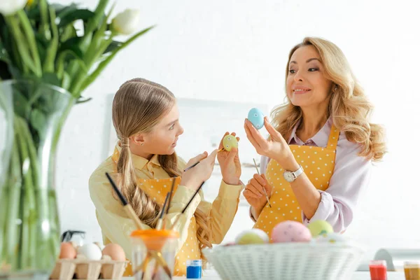 Selective focus of happy mother and daughter holding paintbrushes and painted easter eggs — Stock Photo