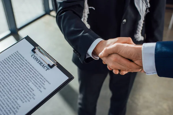 Vista recortada del contrato y los empresarios estrechando la mano en la reunión - foto de stock