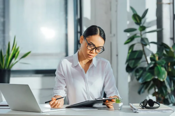 Asian translator in eyeglasses working online with laptop and documents in office — Stock Photo