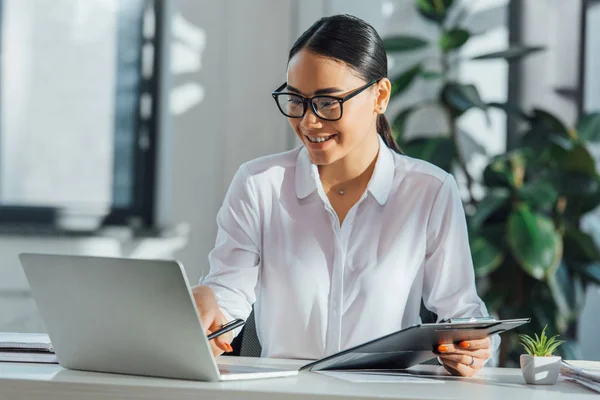 Lächelnder asiatischer Übersetzer mit Brille, der online mit Laptop und Dokumenten arbeitet — Stockfoto