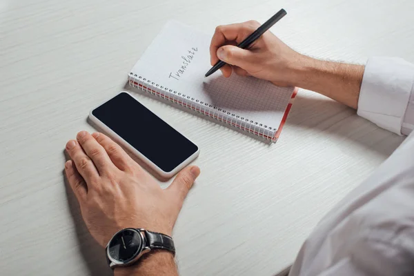 Cropped view of translator writing in notepad with smartphone with blank screen — Stock Photo