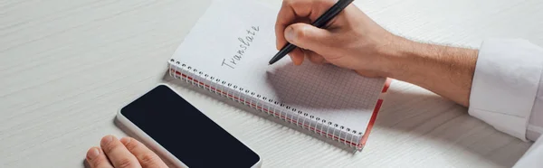 Panoramic shot of male translator writing in notepad with smartphone with blank screen — Stock Photo