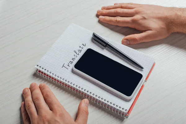 Vista recortada del traductor masculino con bloc de notas, bolígrafo y smartphone con pantalla en blanco - foto de stock