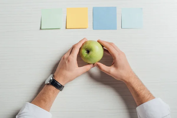 Vue recadrée d'un homme d'affaires tenant une pomme sur une table avec des autocollants vides — Photo de stock