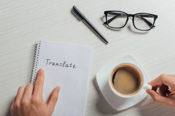 Cropped view of translator with notepad, pen, eyeglasses and cup of coffee — Stock Photo