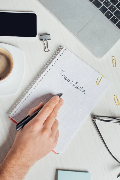 Vista parcial del traductor que trabaja con bloc de notas, laptop y smartphone en el lugar de trabajo con una taza de café - foto de stock