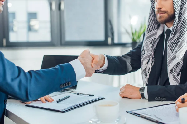 Vista recortada de empresarios multiétnicos estrechando la mano y teniendo acuerdo en la reunión - foto de stock