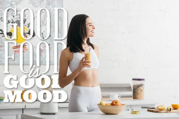 Atractiva deportista en forma sonriendo y sosteniendo vaso de batido cerca de buena comida es ilustración de buen humor - foto de stock