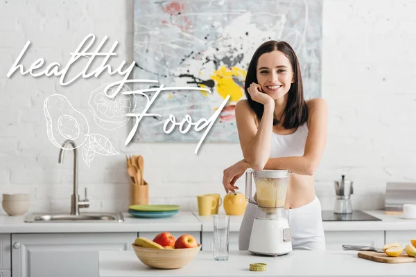 Slim sportiva sorridente alla macchina fotografica mentre prepara frullato vicino a metro nastro sul tavolo della cucina e illustrazione del cibo sano — Foto stock