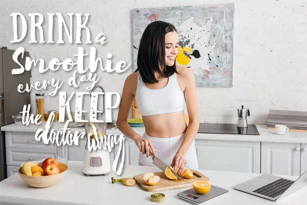 Deportista sonriente en forma mirando a la computadora portátil mientras corta frutas cerca de la licuadora en la mesa de la cocina, bebe un batido todos los días y mantén al médico alejado de la ilustración — Stock Photo