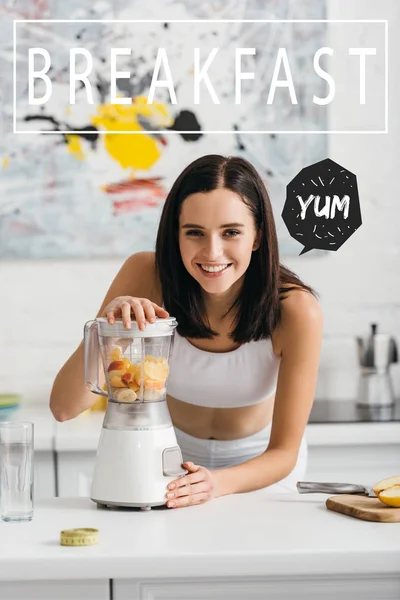 Selective focus of attractive sportswoman smiling at camera while preparing smoothie in blender near breakfast illustration — Stock Photo