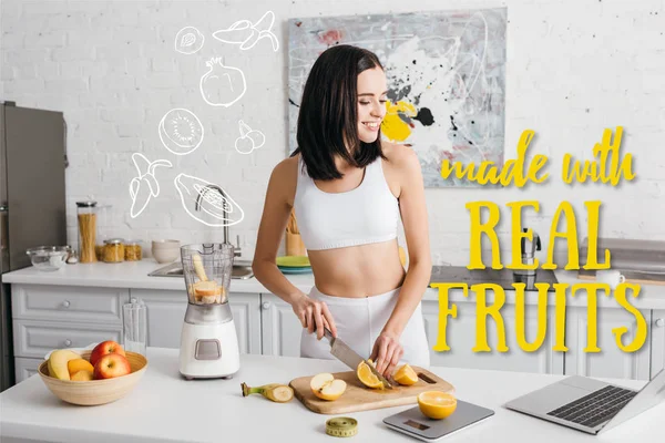 Sorrindo sportswoman ajuste olhando para laptop ao cortar frutas perto do liquidificador na mesa da cozinha, feito com ilustração de frutas reais — Fotografia de Stock