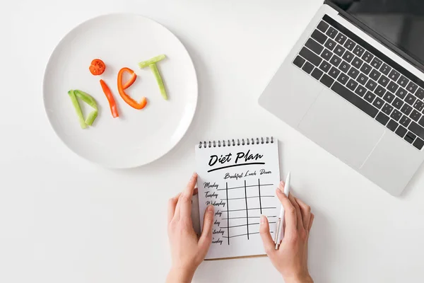 Vista dall'alto della donna che tiene penna e notebook con piano di dieta vicino alla dieta lettering da fette vegetali su piatto e laptop su sfondo bianco — Foto stock